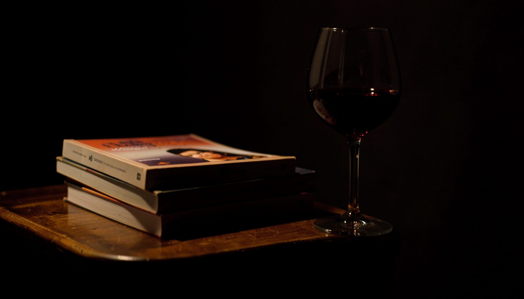 A dimly lit stack of paperback romance novels on a wooden side table beside a half-full wine glass, deep burgundy and amber tones against near-black shadows