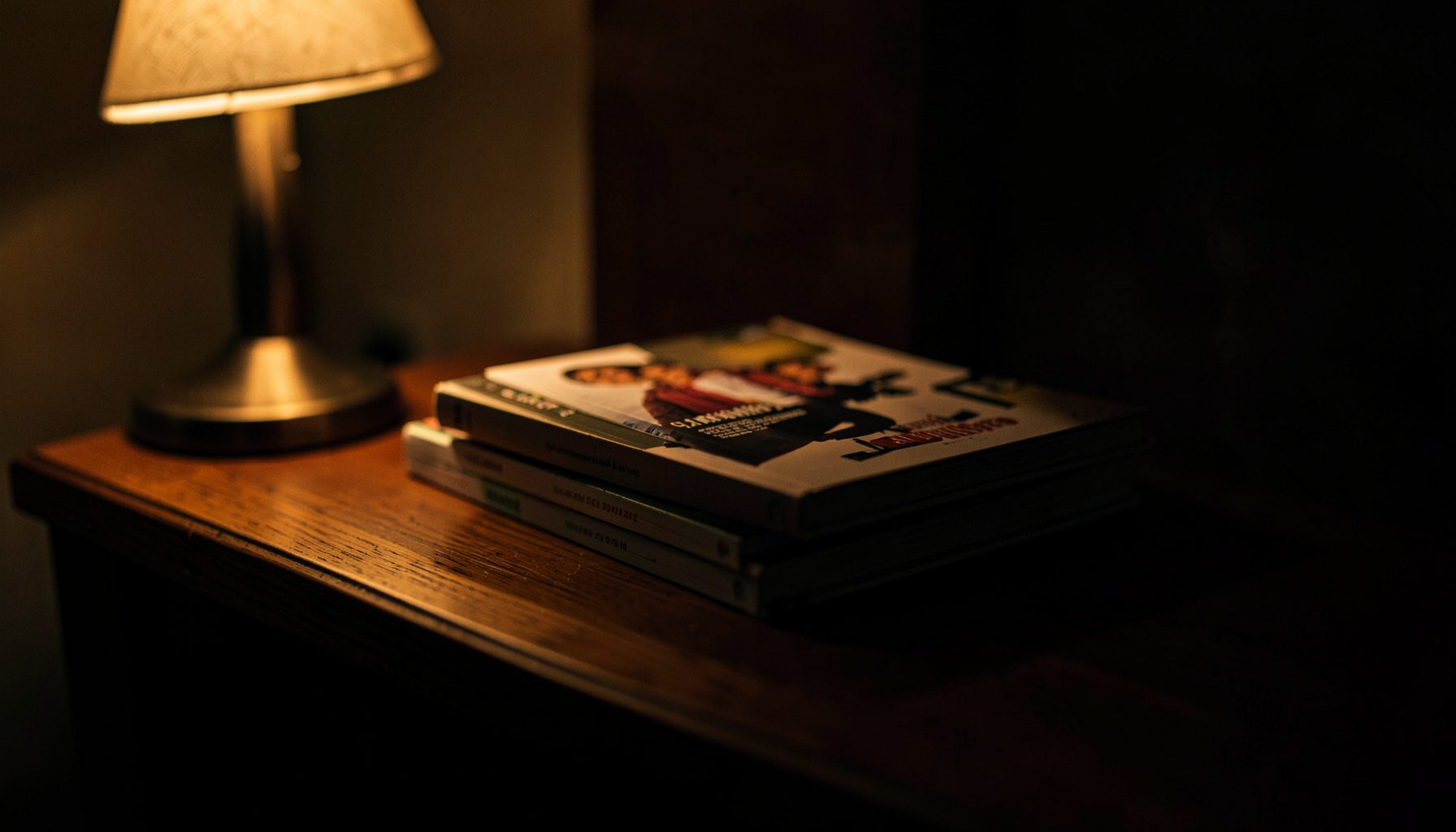 A dimly lit stack of young adult paperbacks on a wooden nightstand, warm amber glow from a bedside lamp, moody and intimate
