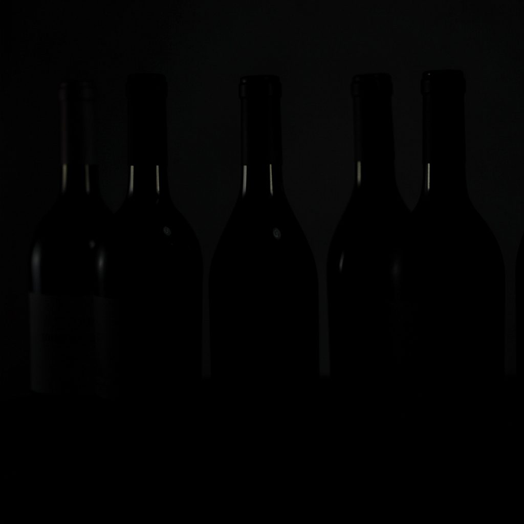 Several wine bottles in silhouette against a dark background, their shapes barely visible in the low-key lighting, with soft highlights on the glass shoulders.