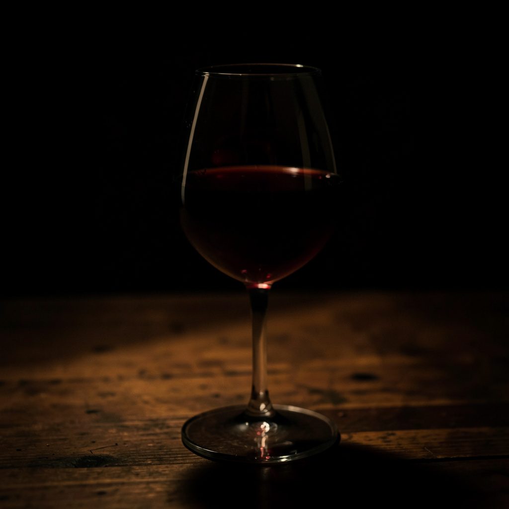 A dark, moody photograph of a half-full wine glass on a wooden table, deep burgundy liquid catching low amber light, with deep shadows pooling around the base.