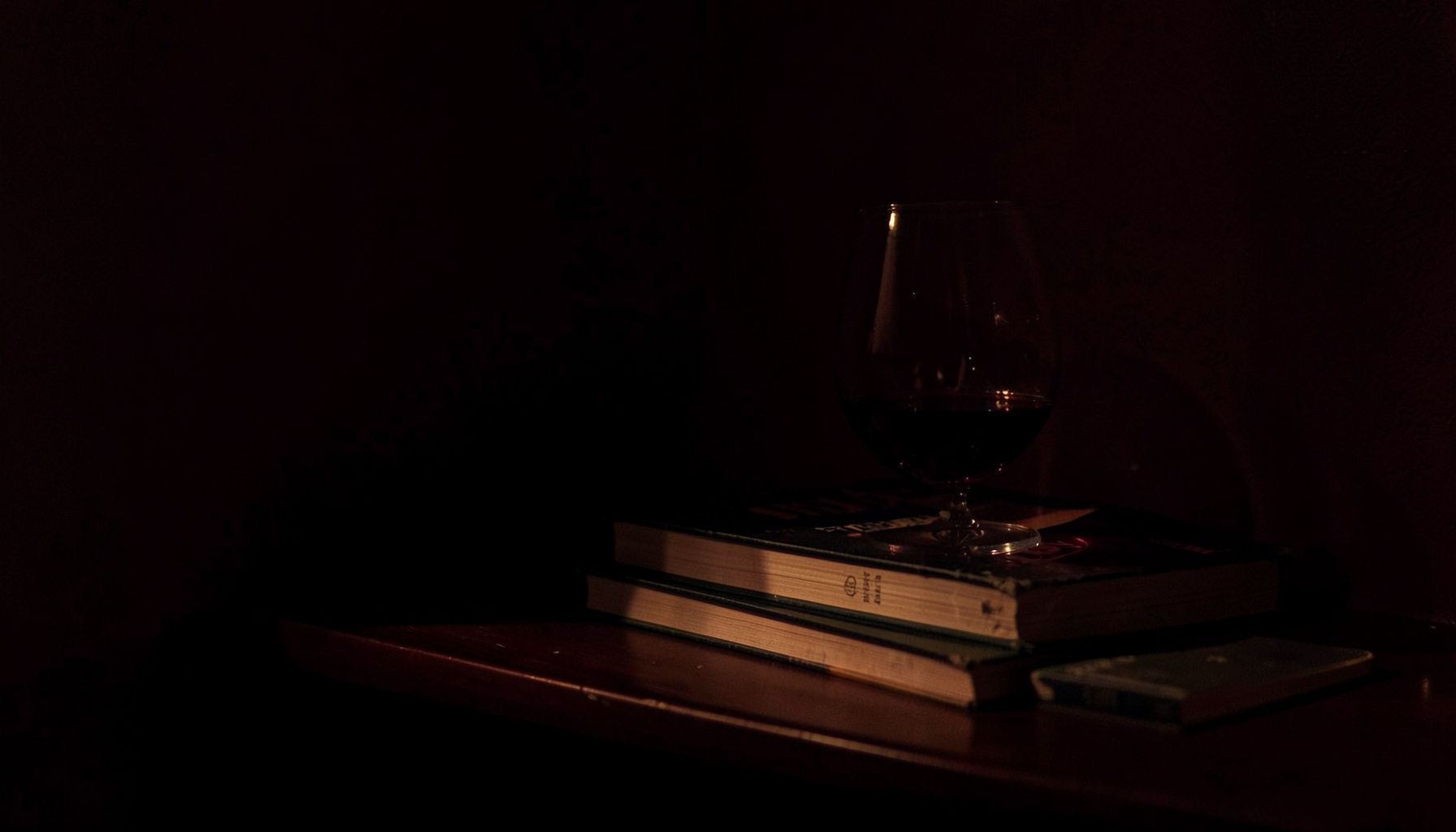 A dimly lit reading nook with a half-empty glass of red wine beside a stack of well-worn paperback novels, deep burgundy and shadow tones